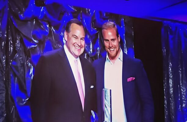 Joe Shew on stage with another man, lit in blue at an awards presentation